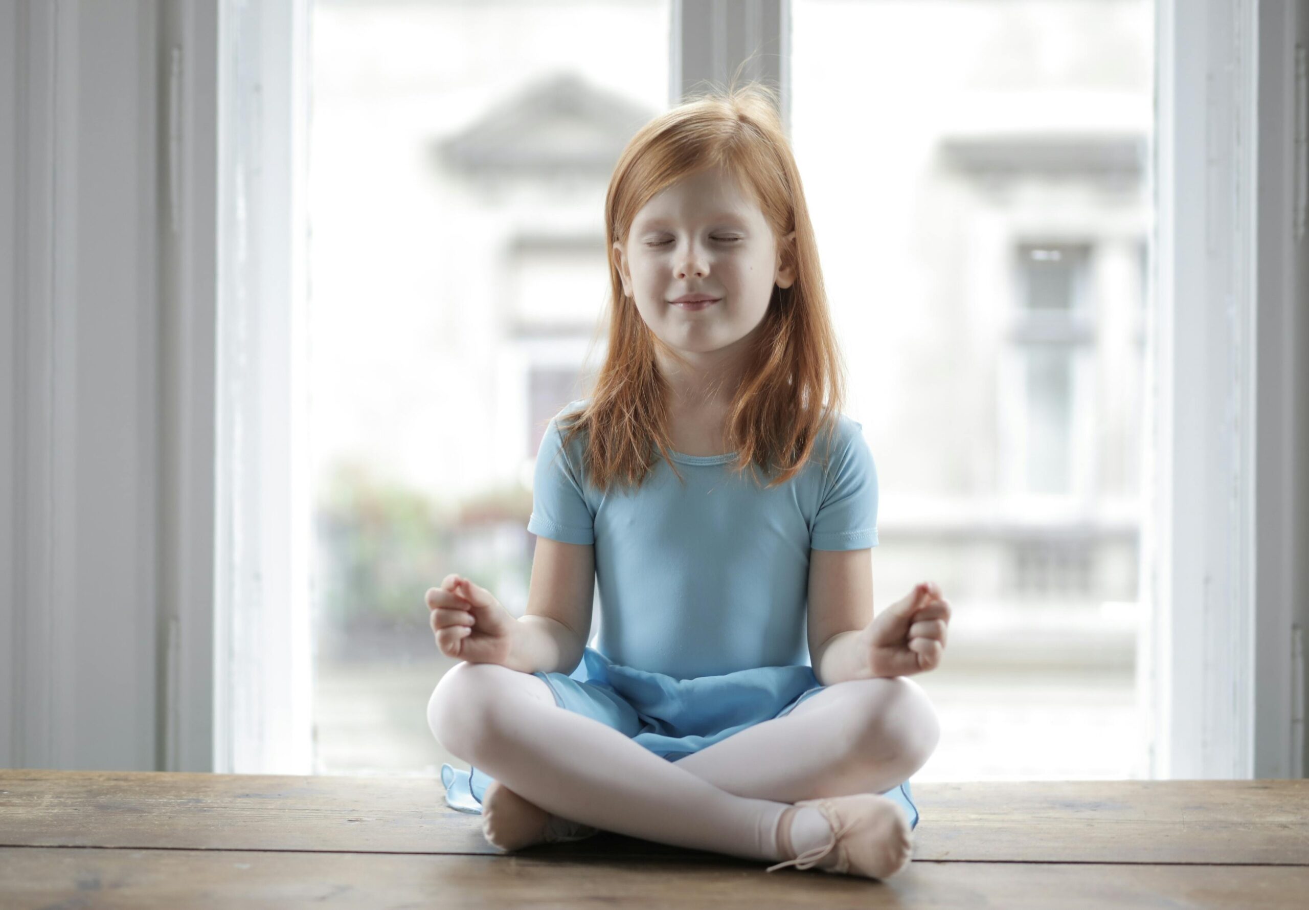 A young girl in a blue dress meditates indoors with serene expression, embracing mindfulness.